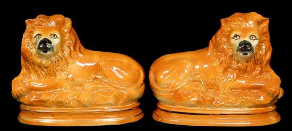 Pair of Staffordshire reclining lions, orange-brown in hue, with off-white faces and gilt and black detailing, reclining on rocky su...